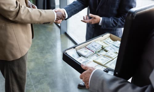 Two businessmen shaking hands while a third person holds an open briefcase full of money.