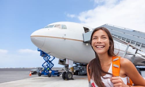 A female tourist on an airport runway carrying a backpack and smiling after having disembarked an airplane.