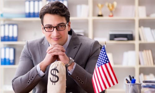 A businessman seated at a table with a bag of money and an American flag.