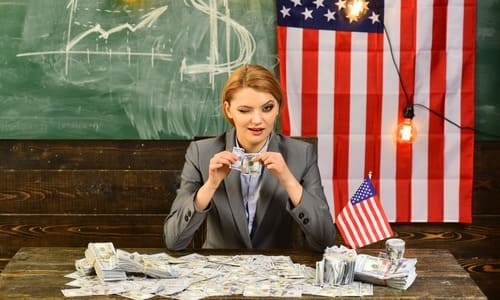 A female CEO seated at a table with the American flag and money laid out and a chalkboard in the background.