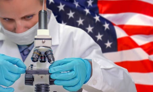 A female scientist looking through a microscope while the American flag waves in the background behind her.