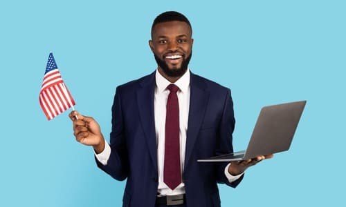 A black businessman smiling and holding a laptop in the left hand, and an American flag in the right.