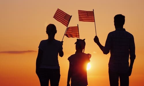 A silhouette of a family of three against the sunset, holding up handheld American flags.