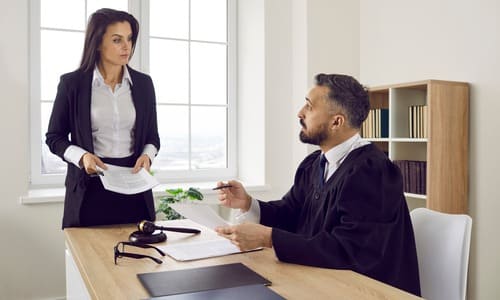 A female lawyer approaching a judge at his desk to hand him papers.