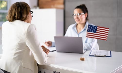 A female lawyer seated at her desk with a laptop and American flag, speaking with a client.