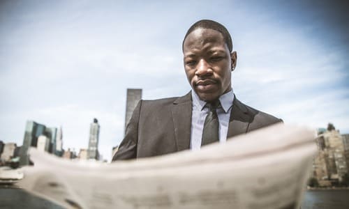 A black businessman with a serious look on his face, reading the newspaper with the New York skyline in the background.