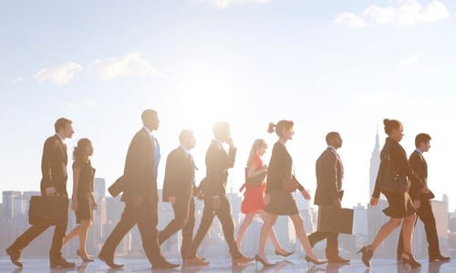 A group of businessmen walking left to right in front of the New York City skyline in the background.