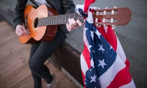 A female musician holding an acoustic guitar with the American flag tied to its headstock.
