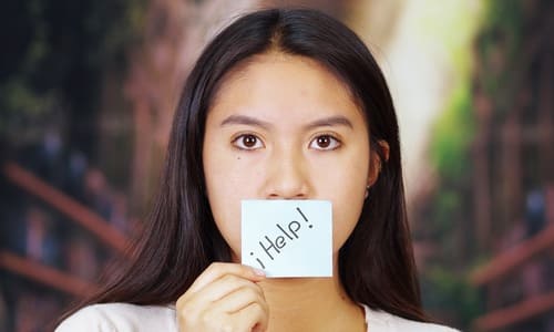 A Southeast Asian woman holding up a piece of paper with the word help written on it up to her mouth.
