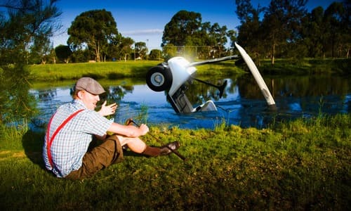 A frustrated golfer seated at the shore of a water hazard on a golf course, with a golf cart sinking in the water.