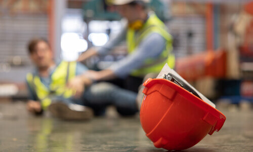 A safety helmet on the ground, with a construction worker in the background tending to injured colleague after an accident.