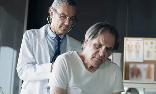 A physician checking the breathing of a patient with a stethoscope against his back.