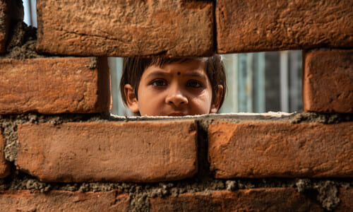 A young Indian girl looking out through a gap in a red brick wall.