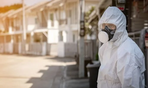 A man in a contamination suit standing in against the background of an empty suburban neighborhood.