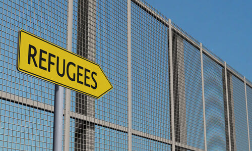 A welded wire mesh fence with a yellow sign labeled "refugees" pointing right.