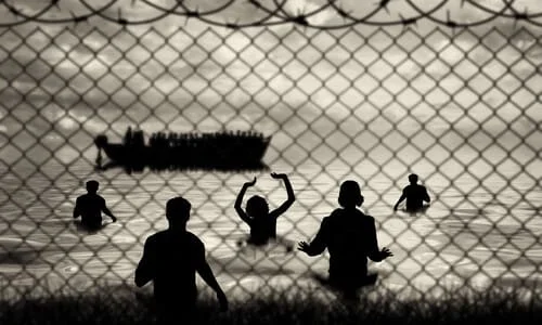 A concept image of silhouetted refugees on a boat arriving at a shore behind wire fence.
