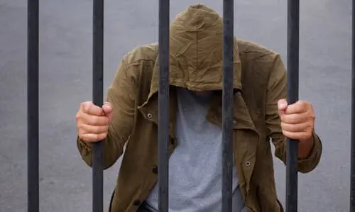 A photo of a hooded figure, presumably a scammer, behind jail bars.