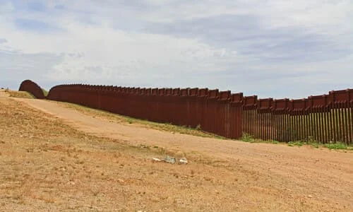 A section of fence along the US-Mexico border next to a dirt road on a hill.