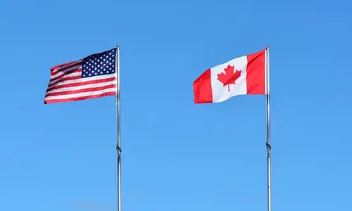 The American and Canadian national flags blowing in the wind against the daytime sky.