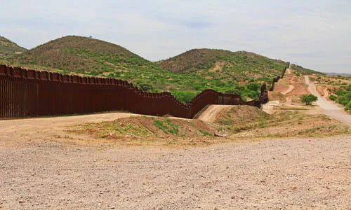 A section of the US-Mexico border fence running alongside a Mexican dirt road over a hill.