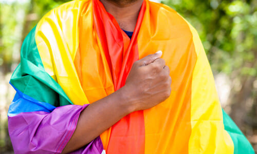 An elderly Indian man praying with a pride flag draped around his shoulders.