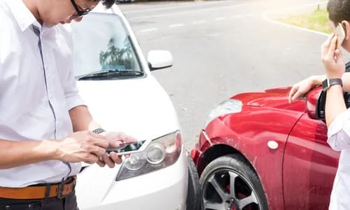 Two male drivers on the phone with presumably their insurance providers after a head-on collision between their cars.