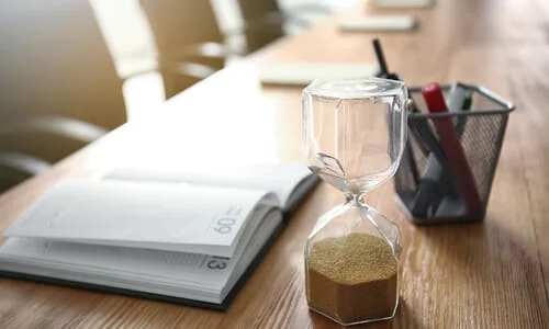 A calendar planner, pen holder, and an hourglass on top of a wooden desk.