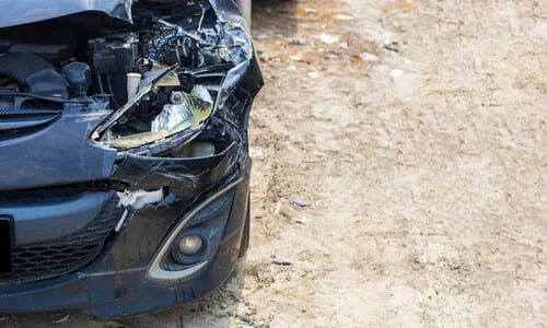 A black car with a broken front bumper and headlight on a city road after an accident.