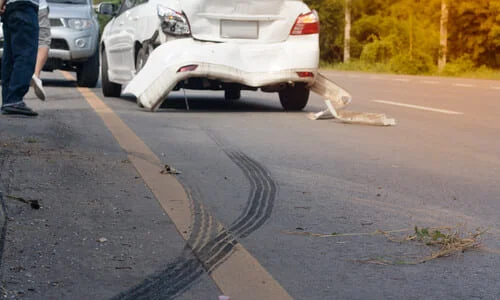 A white car with a torn rear bumper in the middle of the road after an accident.