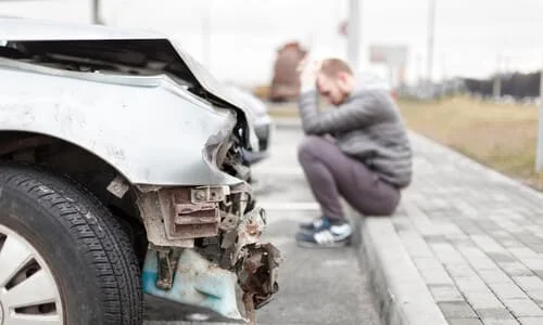 A man sitting on a sidewalk with his hands on his head after crashing his car.