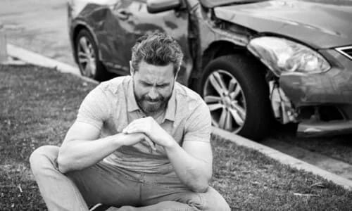 A black-and-white image of a man sitting on a lawn in front of a crashed car