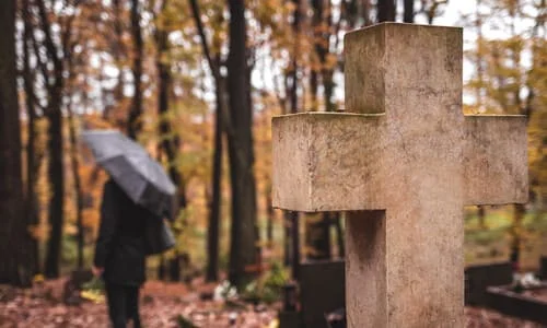 A person in black clothes and with a black umbrella walking in the distance away from a tombstone in the foreground.