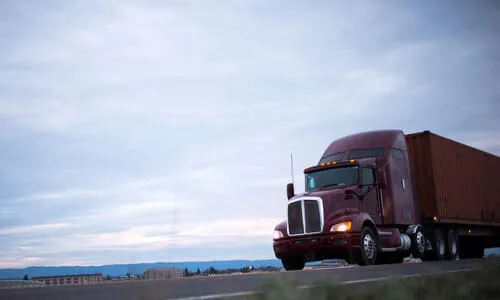 A maroon semi truck driving on a highway road with its trailer in the early evening.